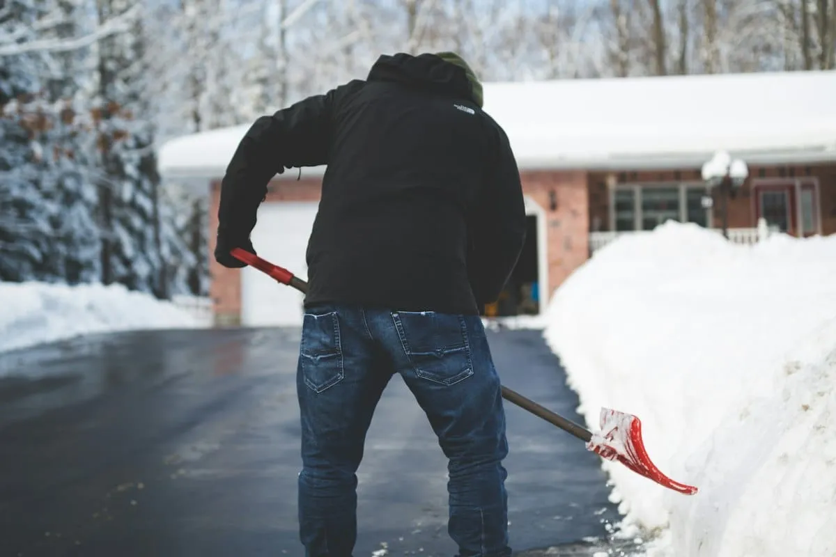 A person shoveling a snow covered driveway in front of a brick house.