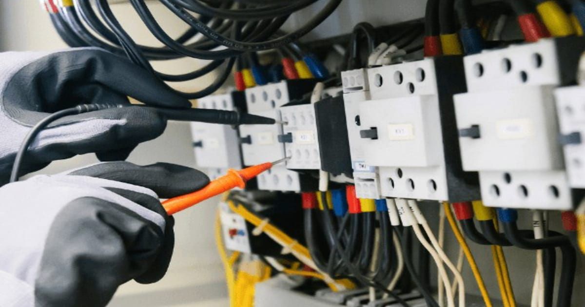 A gloved electrician testing terminals inside an electrical panel with a multimeter probe.