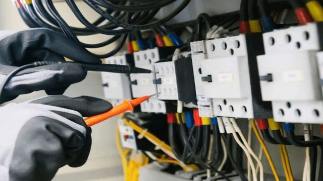 A gloved electrician testing terminals inside an electrical panel with a multimeter probe.